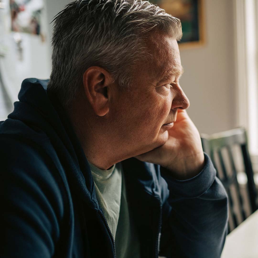 Mandlig pårørende til kræftsyg partner sidder tankefuldt ved køkkenbordet.