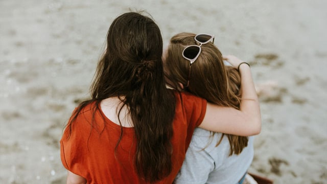 To teenagere, piger, set bagfra på en strand. De holder om hinanden og søger trøst og støtte, når deres mor har brystkræft.