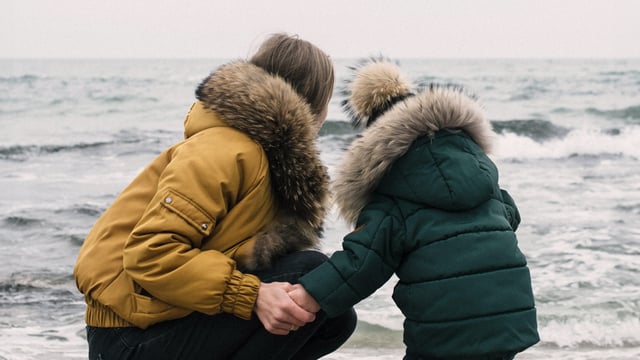 Brystkræftramt kvinde holder sit barn i hånden. De har vinterjakker på og sidder på hug ved vandkanten og ser ud over havet. Illustrationsfoto