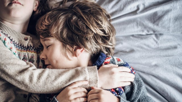 Nærbillede af to børn. En pige der holder om sin bror. De ligger i sengen. Begge har de striktrøjer på. Drengen lukker øjnene og ligger tæt op ad sin søster. Illustrationsfoto