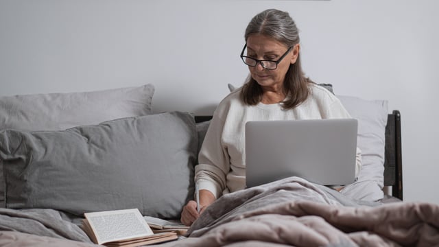 Brystkræftramt kvinde sidder i sengen og arbejder på sin computer. Hun har bøger liggende ved siden af sig og tager notater. Illustrationsfoto