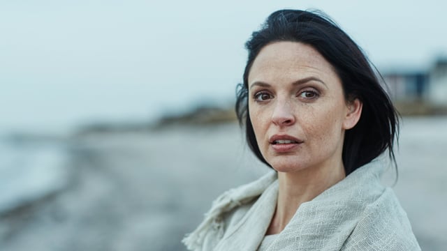 Mørkhåret kvinde med brystkræft med spredning står på stranden og ser ind i kameraet. Illustrationsfoto