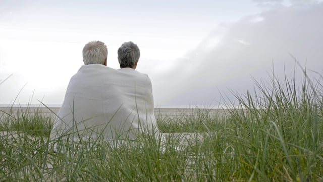 Ældre par, hvor kvinden er ramt af brystkræft, sidder sammen under et tæppe på stranden og ser ud over havet. Vi ser dem bagfra. Illustrationsfoto