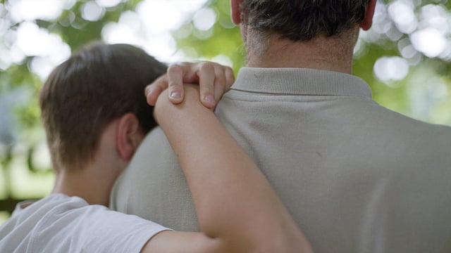 Nærbillede af en søn, der hænger på sin fars skulder og taler om hvad brystkræft med spredning er. Vi ser dem bagfra. Illustrationsfoto